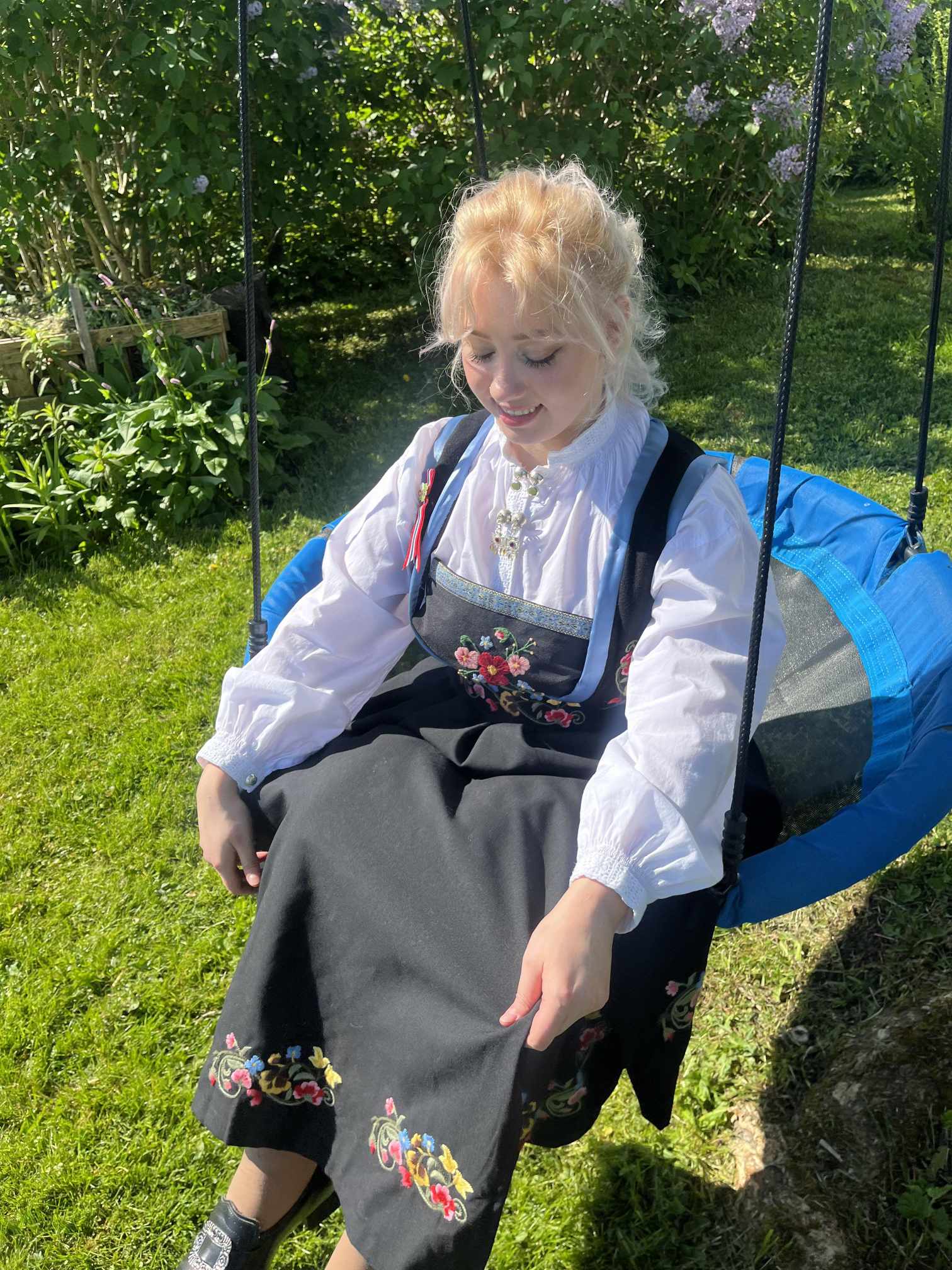 A picture of a young woman dressed in traditional norwegian dress, bunad, sitting on a swing outside.